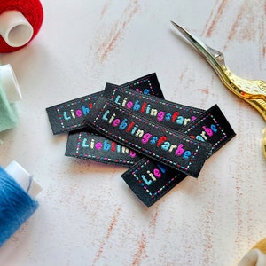 Może przedstawiać: Zbliżenie na kilka czarnych etykiet tekstylnych z wyszytym tekstem "Lieblingsfarbe" kolorową nicią. Etykiety są ułożone jedna na drugiej i mają białe tło.