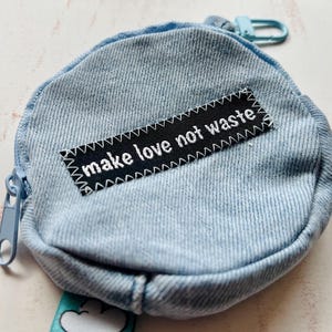 Op de afbeelding: Een lichtblauwe denim munttas met ritssluiting en een kleine karabijnhaak. De tas heeft een zwart label met de witte tekst "make love not waste".