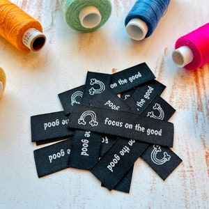 Może przedstawiać: Zbliżenie na stos czarnych etykiet tekstylnych z białym tekstem, który brzmi "focus on the good" i "on the good". Etykiety są ułożone w przypadkowym wzorze, a niektóre z nich mają grafikę tęczy.
