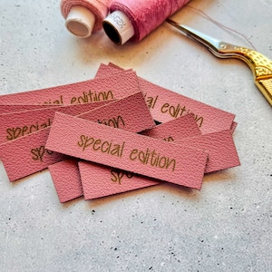 Könnte beinhalten: Ein Stapel rosa Leder-Etiketten mit dem Text "Special Edition" in goldener Schrift.