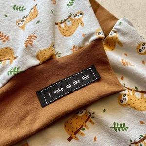 Peut inclure: Un tissu brun et blanc avec un motif de paresseux et le texte "I woke up like this" sur une étiquette noire.