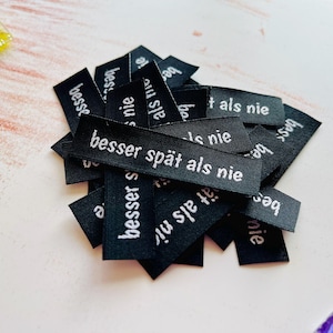 Pode incluir: Um monte de etiquetas de tecido pretas com texto branco que diz "besser spät als nie", que se traduz como "é melhor tarde do que nunca".