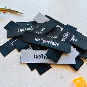 Könnte beinhalten: Ein Stapel schwarzer und weißer Stoff-Etiketten mit dem aufgedruckten Text "un*perfekt". Die Etiketten sind in zufälliger Reihenfolge angeordnet und einige überlappen sich.
