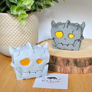 Peut inclure: Deux figurines en pierre de style 3D imprimées en gris avec des yeux jaunes. Les figurines sont posées sur une base en bois.
