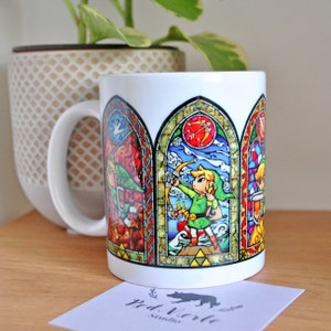 Könnte beinhalten: Weiße Keramiktasse mit einem Buntglasdesign, das eine grün gekleidete Figur mit Pfeil und Bogen zeigt. Das Design ist in drei Abschnitte unterteilt, jeder mit einer anderen Szene. Die Tasse hat einen schwarzen Henkel.