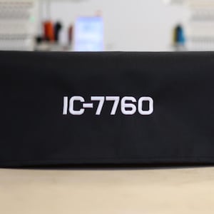 Peut inclure: Deux housses rectangulaires noires avec le texte "IC-7760" brodé en blanc. Les housses sont de tailles différentes et semblent être des étuis de protection. L'arrière-plan est flou, montrant ce qui semble être des machines à broder.