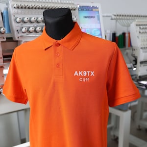 May include: Bright orange polo shirt with short sleeves and a three-button placket. The shirt features the text "AK9TX Cliff" embroidered in white on the chest. The shirt is displayed on a black mannequin.