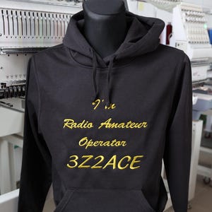 Puede incluir: Sudadera con capucha negra con bolsillo de canguro, exhibida en un maniquí. El texto "I'm Radio Amateur Operator 3Z2ACE" está bordado en hilo dorado en el pecho. Máquina de bordar en el fondo.