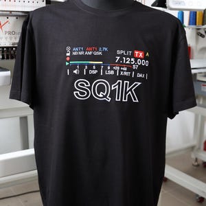 May include: Black t-shirt with white and colourful text. The text includes "ANT1", "SPLIT TX A", "SQ1K", and other technical terms. The shirt is displayed on a black mannequin.