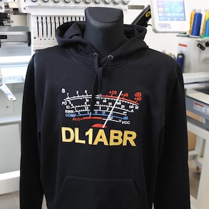Puede incluir: Sudadera con capucha negra con bolsillo de canguro. La parte delantera presenta un gráfico de un medidor con el texto "DL1ABR" en dorado. El medidor tiene marcas para S, PO, IC, SWR, COMP y ALC. La sudadera tiene una capucha con cordón.