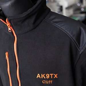 Peut inclure: Veste en polaire noire avec une fermeture éclair orange et un logo brodé noir et orange qui dit "AK9TX Cliff".