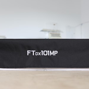 以下が含まれることがあります： 白い文字で「FT Dx101MP」と書かれた黒い長方形のカバー。 カバーは布製で、明るい色の表面に置かれています。 背景はぼやけており、他のオブジェクトが少し見えます。