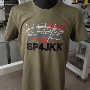 Puede incluir: Una camiseta verde oliva con un gráfico bordado de un medidor de señal de radio con escalas y agujas en blanco, rojo y azul. El texto SP4JKK está bordado en letras negras grandes debajo del gráfico en la parte frontal de la prenda.