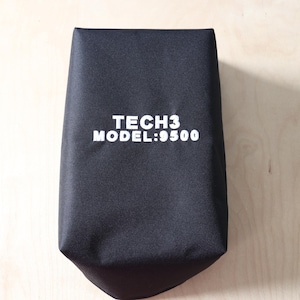 May include: A black, rectangular bag with the text "TECH3 MODEL:9500" printed in white. The bag is made of a textured material and is set against a light wood background.