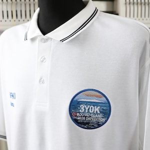 Peut inclure: Polo blanc à col et manches courtes. Le col et les poignets des manches ont des rayures noires et blanches. Un écusson circulaire sur la poitrine porte l'inscription "3Y0K Bouvet Island 2026 DXpedition". Le t-shirt porte également l'inscription "WKI" en bleu.