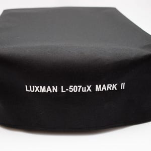 Puede incluir: Una funda negra con el texto "LUXMAN L-507uX MARK II" en blanco. La funda parece estar hecha de un material suave y mate. El artículo es probablemente una funda protectora para un dispositivo electrónico.