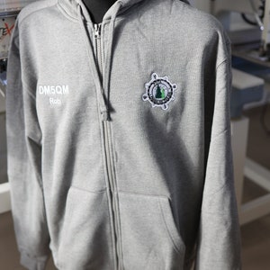 Peut inclure: Sweat-shirt à capuche gris avec fermeture éclair. Le sweat-shirt a un logo brodé blanc avec une boussole et des arbres sur le côté droit de la poitrine. Le texte "DM5QM Rob" est brodé en blanc sur le côté gauche de la poitrine.