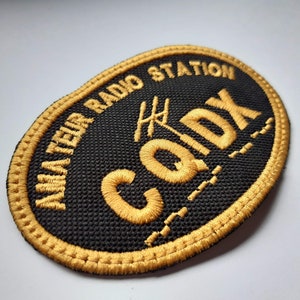 May include: A black embroidered patch with gold stitching. The patch reads "Amateur Radio Station Cody" with an antenna symbol above the name.