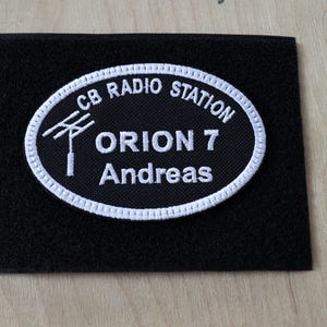 Puede incluir: Parche rectangular negro con un diseño ovalado bordado en blanco. El óvalo dice "CB RADIO STATION" en la parte superior, con un gráfico de torre de radio y el texto "ORION 7 Andreas" en el centro. El parche está sobre una superficie de madera clara.