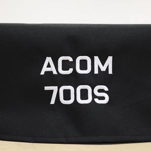 Könnte beinhalten: Schwarze rechteckige Hülle mit dem weißen Text "ACOM 700S" auf der Vorderseite gestickt. Die Hülle scheint aus einem strapazierfähigen, strukturierten Stoff zu bestehen. Der Hintergrund ist einfarbig weiß.