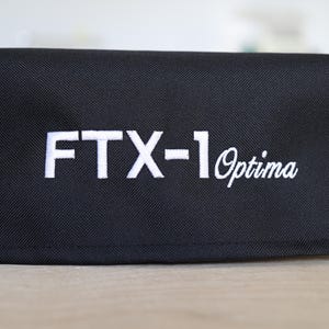 Pode incluir: Estojo retangular preto com o texto bordado "FTX-1 Optima" em branco. O estojo parece ser feito de um tecido durável e texturizado. O fundo está desfocado, focando a atenção no produto.