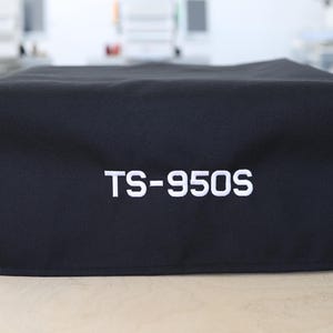 Pode incluir: Uma capa de tecido preto com o texto bordado "TS-950S" em branco. A capa, de forma retangular, é provavelmente projetada para proteger um equipamento. O fundo está desfocado, sugerindo um ambiente interno.