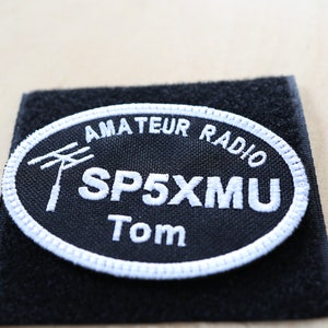 Pode incluir: Um remendo oval preto com costuras brancas. O remendo diz "Amateur Radio SP5XMU Tom" com um símbolo de antena branco.