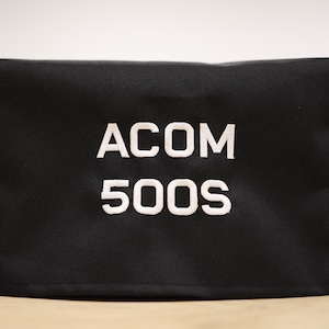 Pode incluir: Capa de tecido preto com texto bordado branco que diz "ACOM 500S".