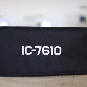 Pode incluir: Capa de tecido preto com texto bordado em branco "IC-7610".