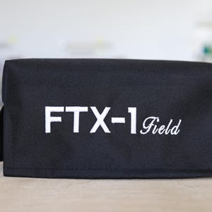 May include: Black rectangular fabric case with the text "FTX-1 Field" embroidered in white. The case is standing upright with two flaps extended, and is on a light-coloured surface.