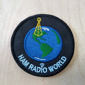 Puede incluir: Parche bordado negro con un globo terráqueo azul y verde, una antena amarilla y el texto "HAM RADIO WORLD".
