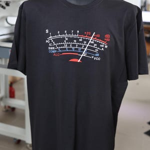Peut inclure: T-shirt noir avec un graphique d'un indicateur avec des marquages rouges, blancs et bleus. L'indicateur affiche des valeurs telles que S, PO, SWR, COMP et ALC. Le t-shirt indique également +20, +40 et +60 dB.