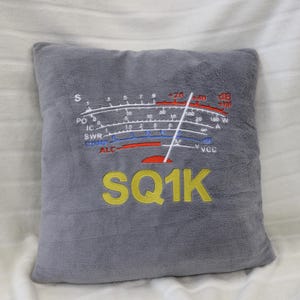 Peut inclure: Un coussin en peluche gris avec un motif de compteur brodé en blanc, rouge et bleu. Le texte "SQ1K" est brodé en jaune. Le coussin est carré et posé sur une surface blanche.