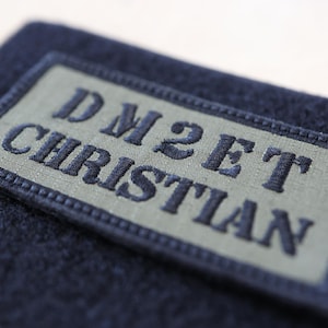 Può includere: Primo piano di un tessuto blu navy con una toppa rettangolare. La toppa è verde chiaro con il testo ricamato "DM2ET CHRISTIAN" in blu navy. La toppa è bordata con cuciture blu navy.
