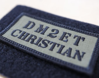 Könnte beinhalten: Nahaufnahme eines marineblauen Stoffes mit einem rechteckigen Aufnäher. Der Aufnäher ist hellgrün mit dem gestickten Text "DM2ET CHRISTIAN" in Marineblau. Der Aufnäher ist mit marineblauen Nähten eingefasst.
