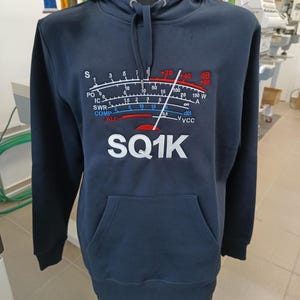 Może przedstawiać: Ciemnoniebieska bluza z kapturem z białym haftem przedstawiającym radiometr z napisem "SQ1K" poniżej.