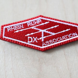Puede incluir: Parche rojo bordado con costuras blancas. El parche tiene forma de hexágono y presenta el texto "Rhein Ruhr DX Association" y un diseño de antena estilizado.