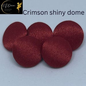 May include: Five circular buttons with a shiny crimson fabric finish and a domed shape. The buttons are arranged in a cluster, displaying a smooth, reflective texture across their surfaces. The text "Crimson shiny dome" is visible above them.
