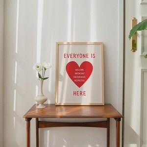 Może przedstawiać: Oprawiony wydruk z napisem "EVERYONE IS HERE" w kolorze czerwonym, z czerwonym sercem pośrodku. Serce zawiera słowa "WELCOME, IMPORTANT, ENCOURAGED, RESPECTED". Wydruk znajduje się na drewnianym stole z wazonem białych kwiat&oacute;w.
