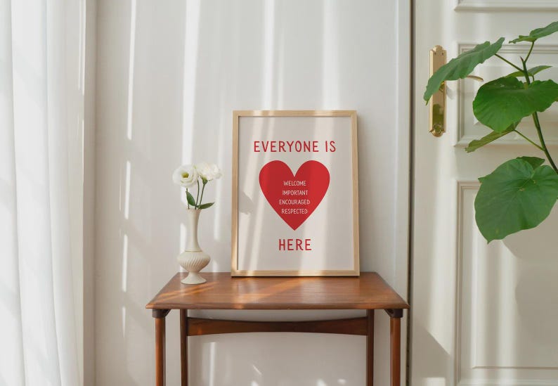 Może przedstawiać: Oprawiony wydruk z napisem "EVERYONE IS HERE" w kolorze czerwonym, z czerwonym sercem pośrodku. Serce zawiera słowa "WELCOME, IMPORTANT, ENCOURAGED, RESPECTED". Wydruk znajduje się na drewnianym stole z wazonem białych kwiat&oacute;w.