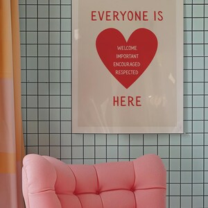 Może przedstawiać: Oprawiony plakat ze słowami "EVERYONE IS HERE" na czerwono, z czerwoną grafiką serca zawierającą słowa "WELCOME, IMPORTANT, ENCOURAGED, RESPECTED". Plakat wisi na jasnoniebieskiej kafelkowej ścianie. R&oacute;żowy fotel znajduje się na pierwszym planie.