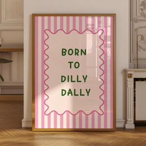 May include: Framed art print with a light brown wooden frame. The artwork features a pink and white striped background with a pink wavy border. The text "BORN TO DILLY DALLY" is written in green, centered within the pink border.