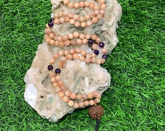 May include: A mala or prayer beads made of pink and orange sunstone beads with purple amethyst accents. The mala is displayed on a white crystal cluster and has a wooden bead at the end.