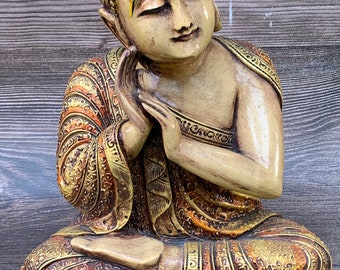May include: A seated Buddha statue with a golden and orange patterned robe. The statue has a serene expression and closed eyes. The statue is made of wood and has a hand resting on the cheek.