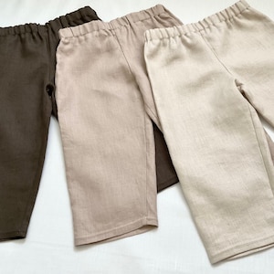 May include: Three pairs of linen trousers in brown, beige, and light beige. The trousers have an elastic waistband and a straight leg.