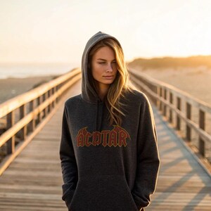 May include: Dark gray hooded sweatshirt with the word "ACOTAR" in a red and gold design. The sweatshirt has a drawstring hood and a kangaroo pocket. The person is standing on a wooden boardwalk.