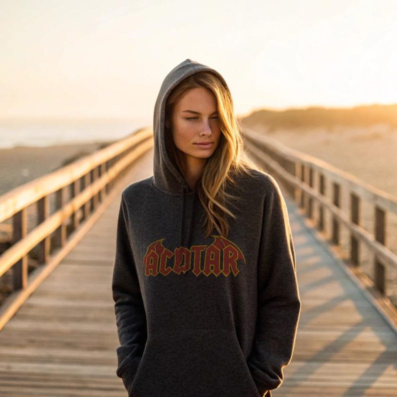 May include: Dark gray hooded sweatshirt with the word "ACOTAR" in a red and gold design. The sweatshirt has a drawstring hood and a kangaroo pocket. The person is standing on a wooden boardwalk.
