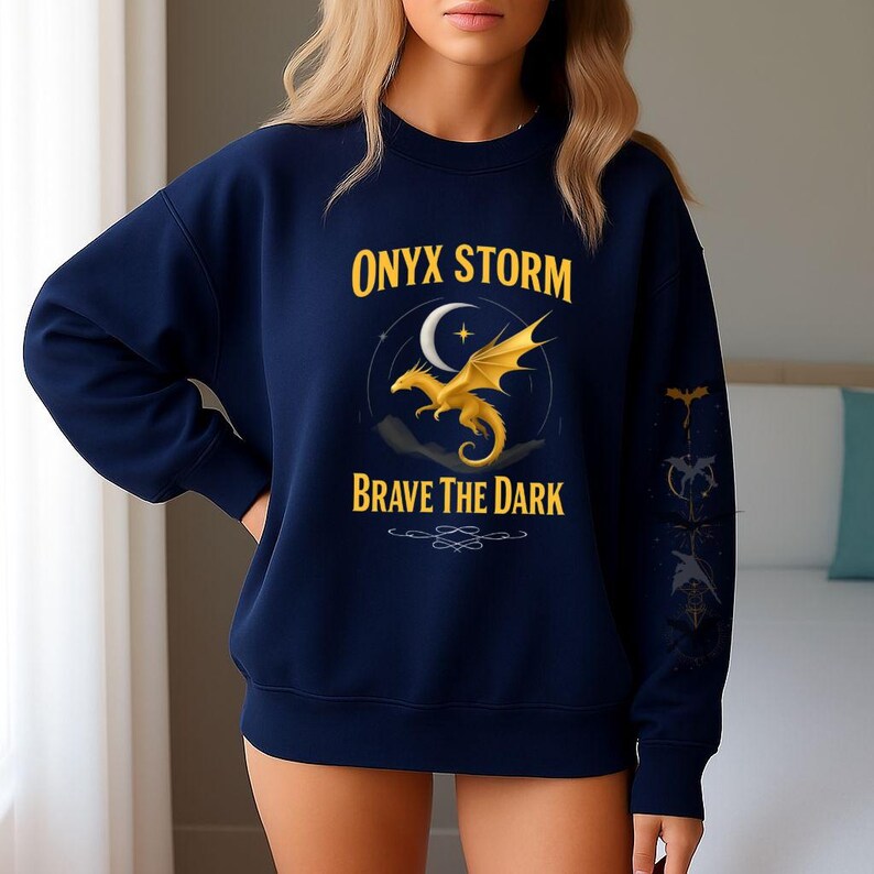 Puede incluir: Sudadera azul marino con el texto "ONYX STORM" sobre un dise&ntilde;o de drag&oacute;n dorado y luna creciente. El texto "BRAVE THE DARK" est&aacute; debajo del drag&oacute;n. Un gr&aacute;fico de drag&oacute;n est&aacute; en la manga derecha.