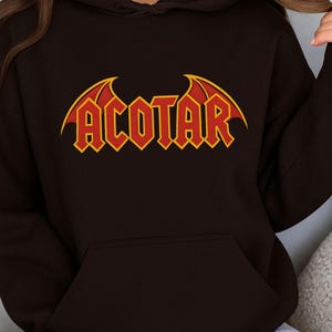 May include: A dark brown hoodie with a front pocket and a hood. The word "ACOTAR" is printed across the chest in red block letters with yellow accents and bat-wing designs.