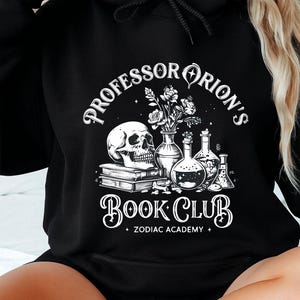 May include: Black hoodie featuring a graphic with the text "Professor Orion's Book Club" and illustrations of a skull, books, flowers, and laboratory glassware. The design includes the words "Zodiac Academy".
